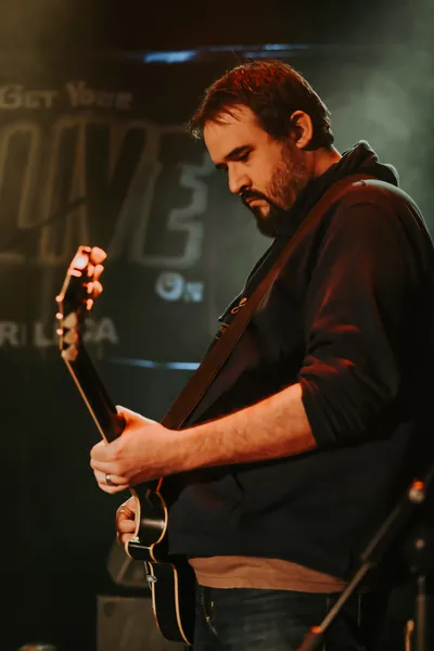 A bearded, hooded hotshot leaning into a well-worn electric guitar, fingers flashing with fierce precision, face fixed in focused intensity, bathed in bold stage light, carving a searing, soaring lead line.
