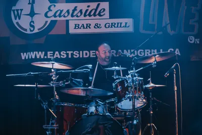 A serious, steady-eyed drummer seated at a scuffed, steel-snared drum kit, sticks set, spine straight, spotlit by subtle stage glow, prepared to pound a precise, pulsing pattern.